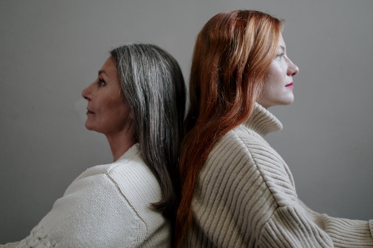 A close-up side view of two women sitting back to back in knitted sweaters, featuring a neutral background.